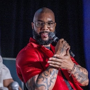 Man with glasses and tattoos wearing a red shirt holding a microphone while speaking on stage